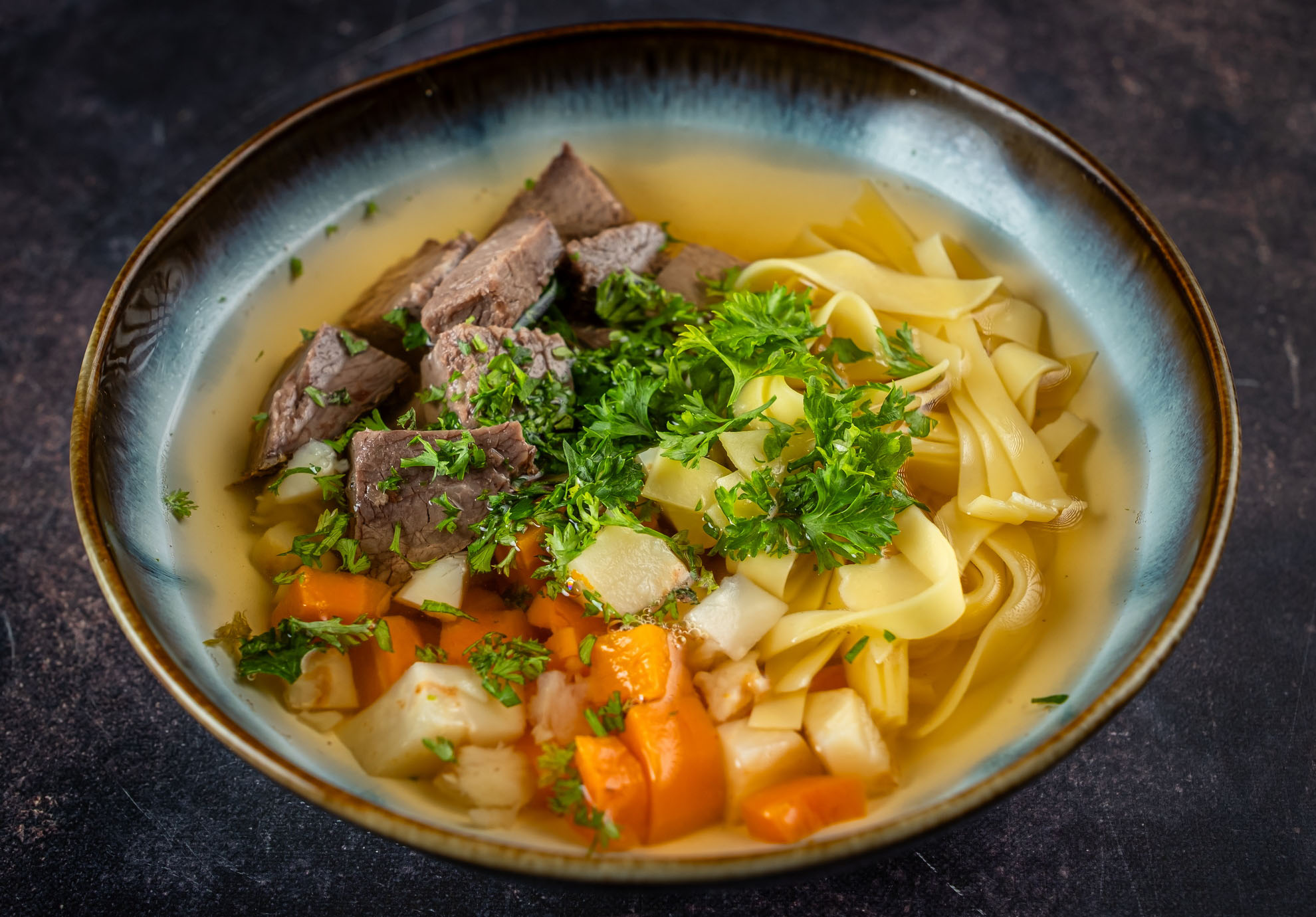 Egy tál kiadós marhahúsos tésztaleves, puha marhahúsdarabokkal, szeletelt sárgarépával, kockára vágott burgonyával, tojásos tésztával és friss petrezselyemmel díszítve, sötét háttér előtt.